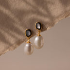 The Midnight Pearl Drop Earrings, featuring gold, pearls, and black accents, rest on a beige textured fabric background and cast soft shadows—a display of timeless jewelry elegance.