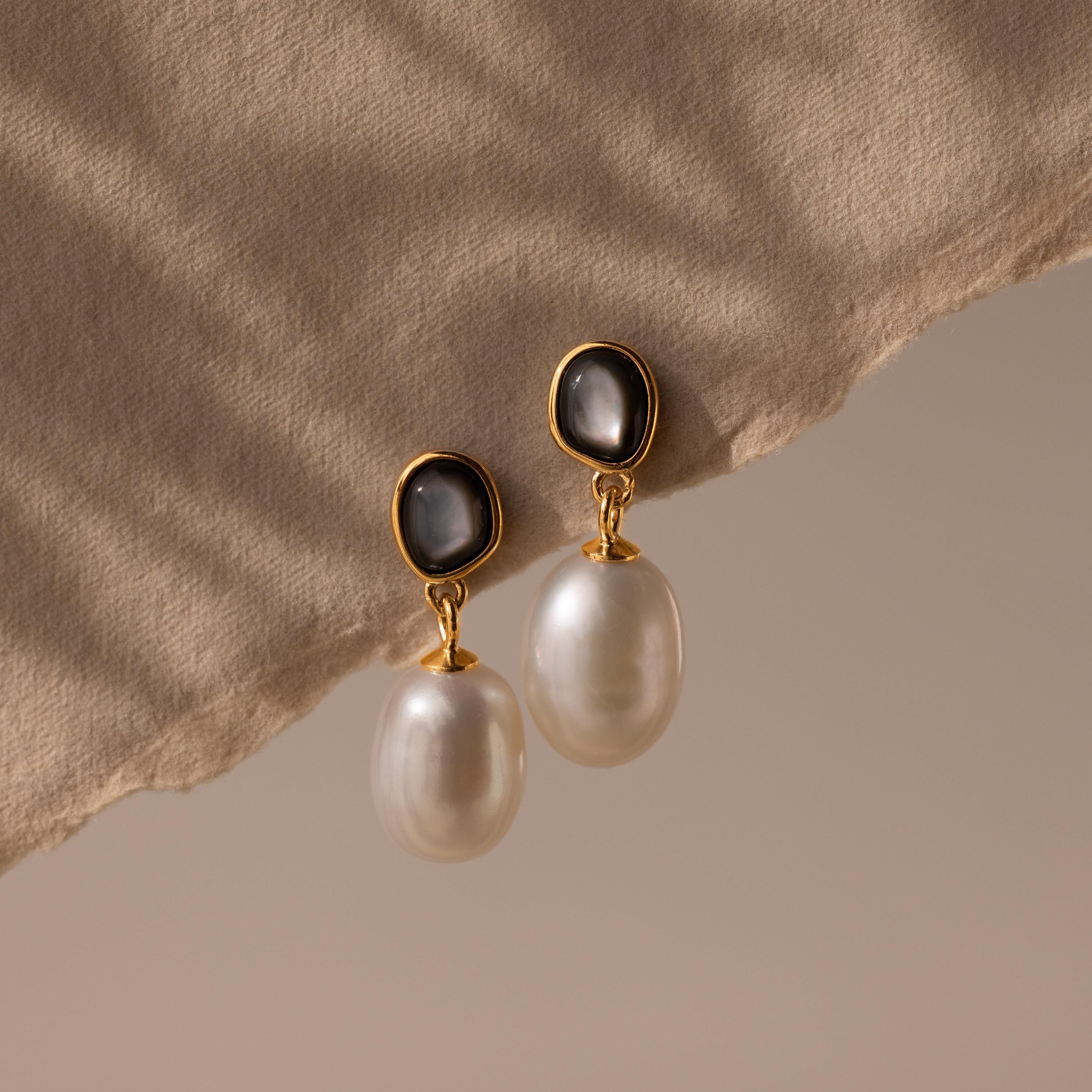 The Midnight Pearl Drop Earrings, featuring gold, pearls, and black accents, rest on a beige textured fabric background and cast soft shadows—a display of timeless jewelry elegance.