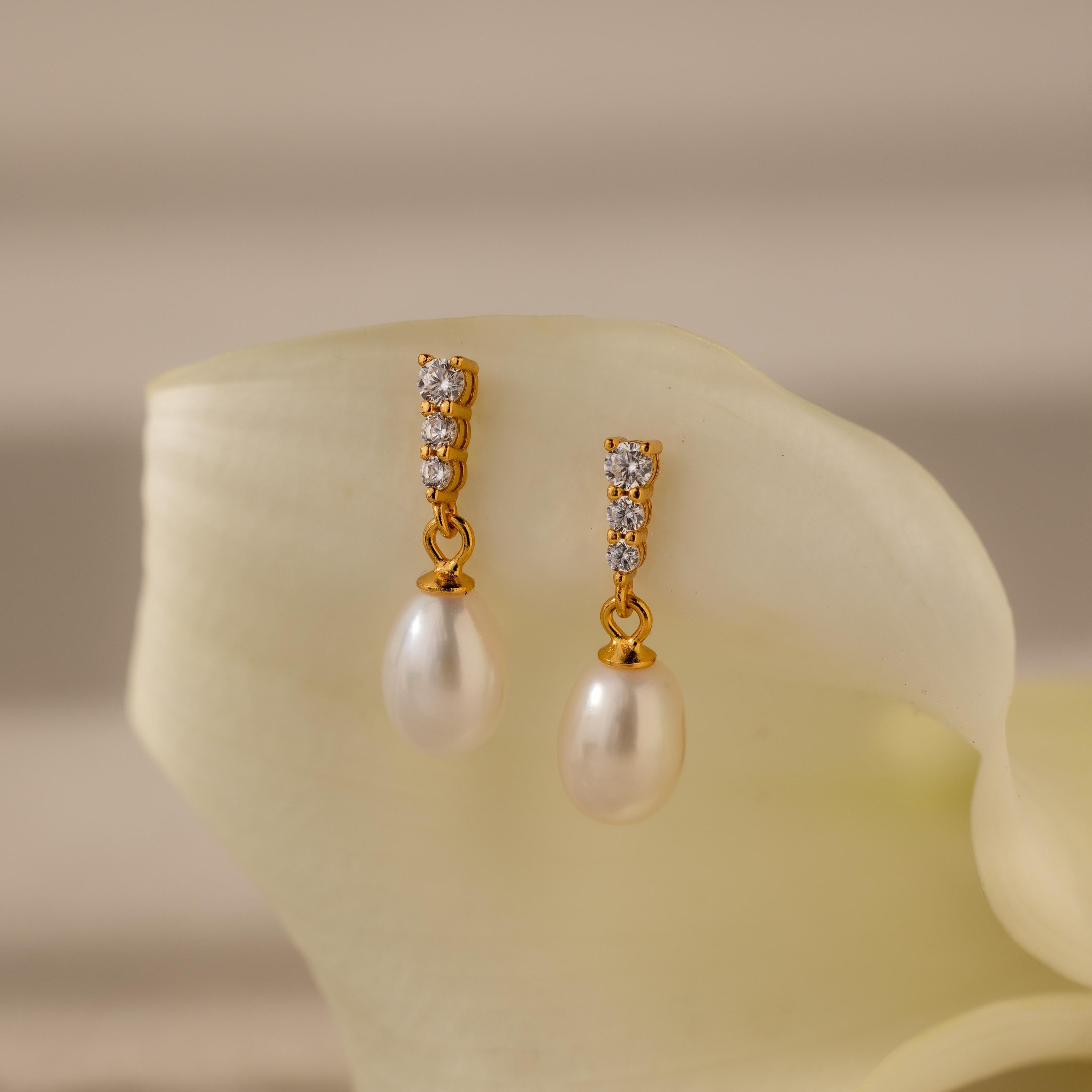 The Diamond Pearl Droplet Earrings in 18K Gold feature delicate pearl drops and sparkling diamonds, beautifully displayed on a white flower petal with a soft background.