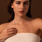 Woman wearing a strapless top and the Tiny Pearl Dot Necklace, gently touching the delicate jewelry, against a brown background.
