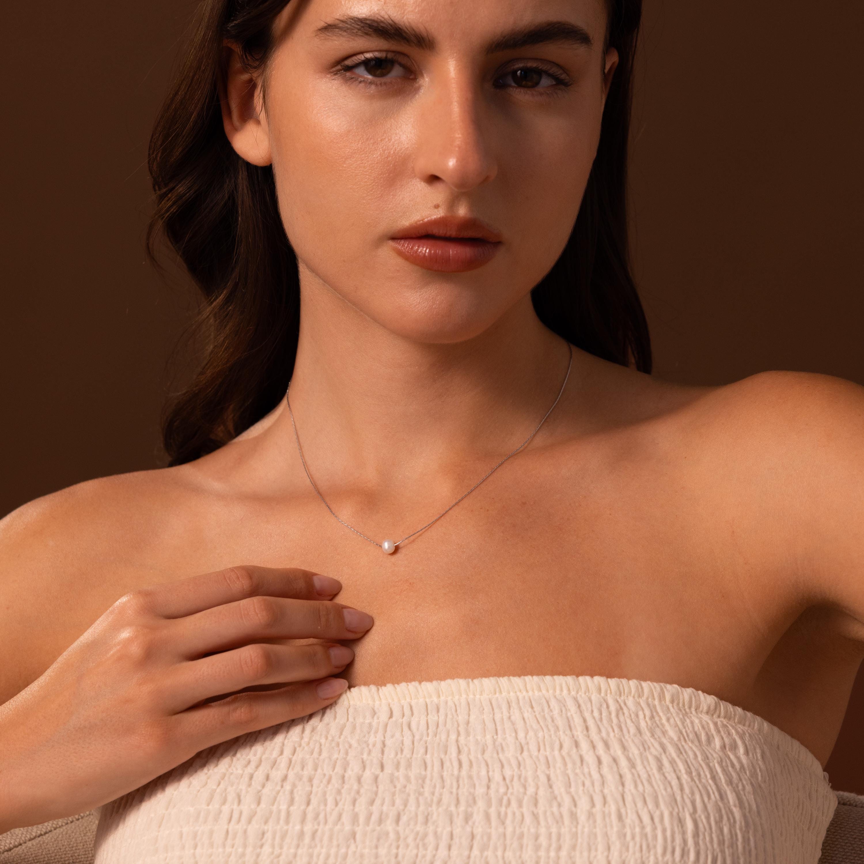 Woman wearing a strapless top and the Tiny Pearl Dot Necklace, gently touching the delicate jewelry, against a brown background.
