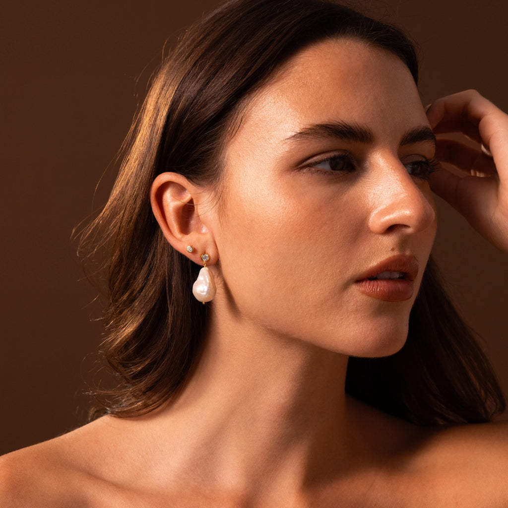Woman with long brown hair wears Diamond Baroque Pearl Studs in 18K Gold, gazing to the side against a brown background.