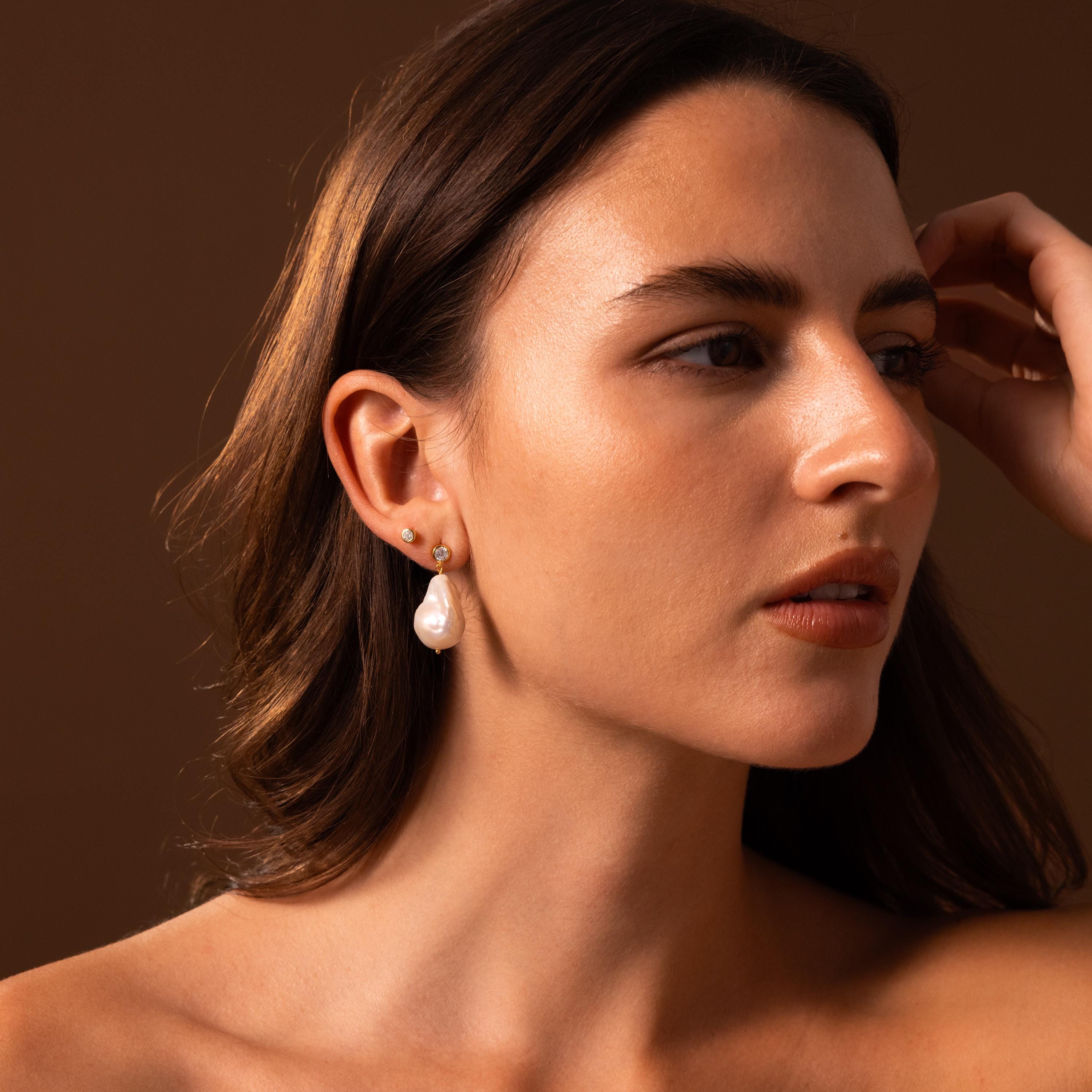 Woman with long brown hair wears Diamond Baroque Pearl Studs in 18K Gold, gazing to the side against a brown background.