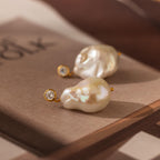 The Diamond Baroque Pearl Studs in 18K Gold feature two organic baroque pearls accented by small round diamonds, creating a stunning piece of jewelry displayed on a book.