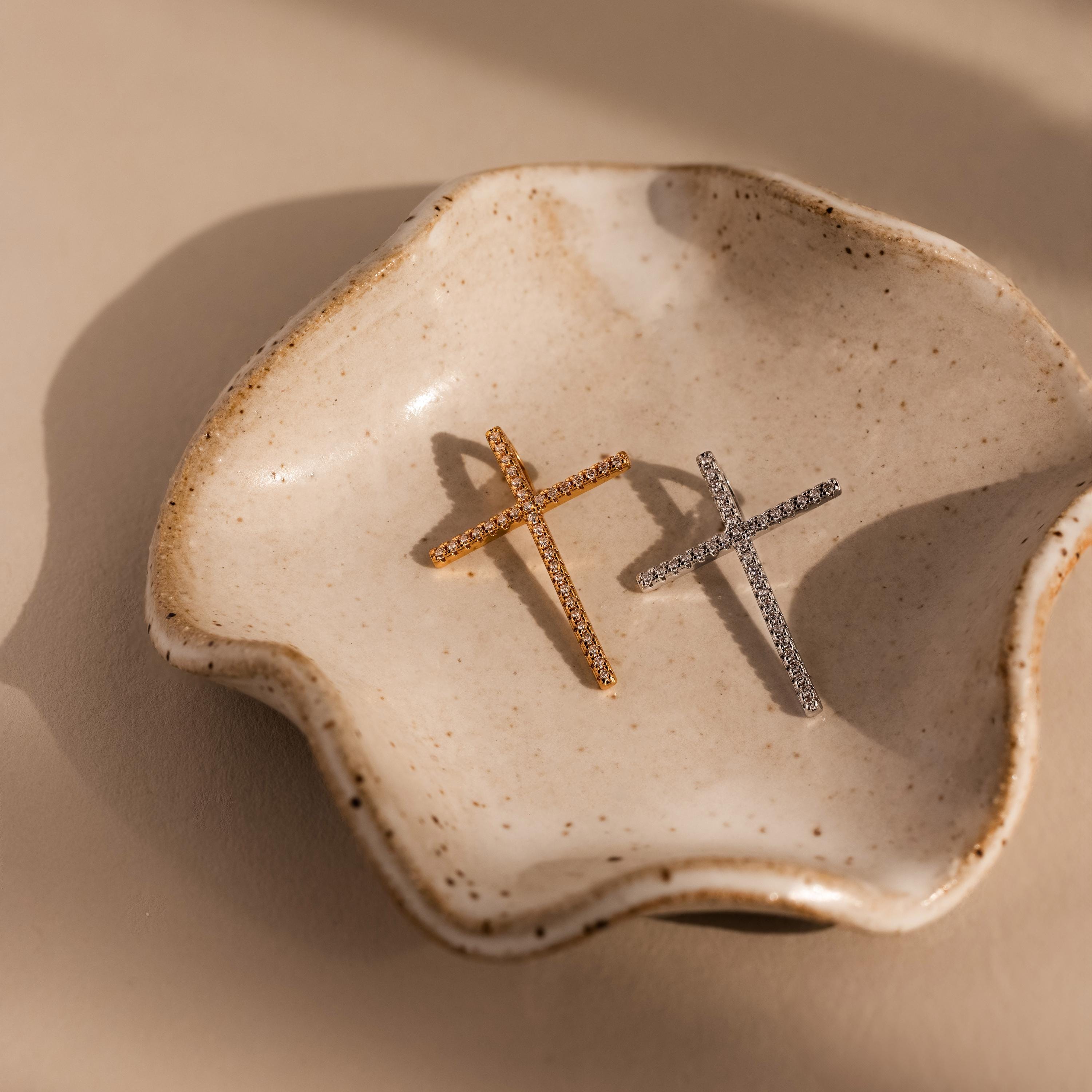 The Skinny Pave Cross Charm, shown in both gold and silver, rests on a wavy ceramic dish under soft lighting—an elegant display of faith jewelry with delicate sparkle.