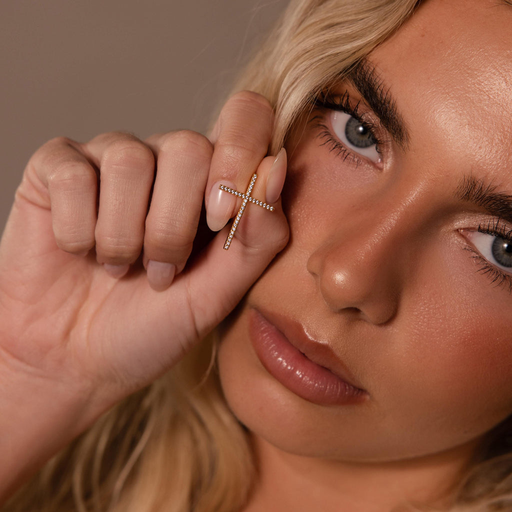 A woman holds the Skinny Pave Cross Charm in 18K Gold earring near her face, highlighting her eyes and natural makeup—an elegant showcase of modern faith jewelry.