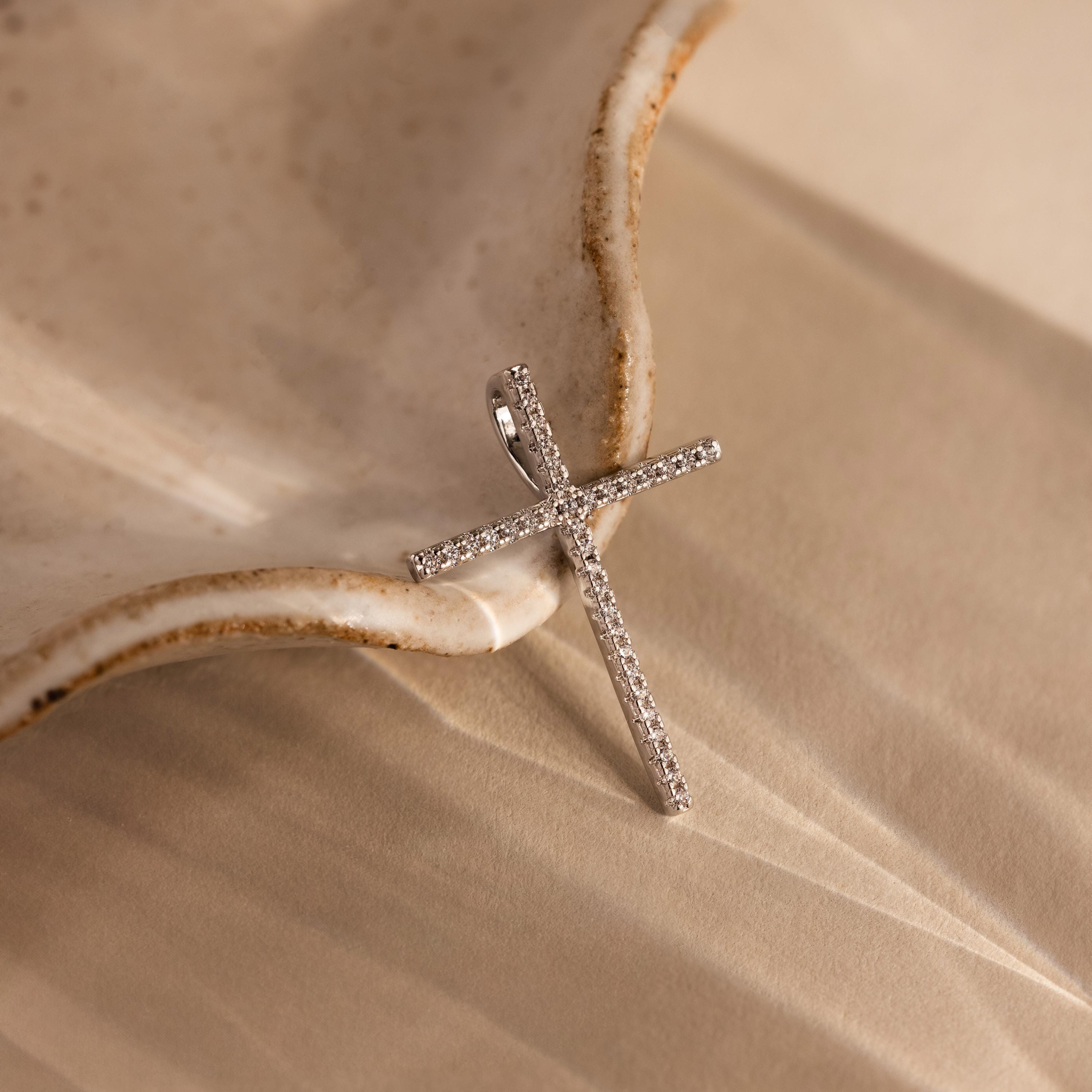 The Skinny Pave Cross Charm in Silver, adorned with small diamonds, rests on a beige textured surface beside a ceramic dish, highlighting the elegance of cross jewelry.