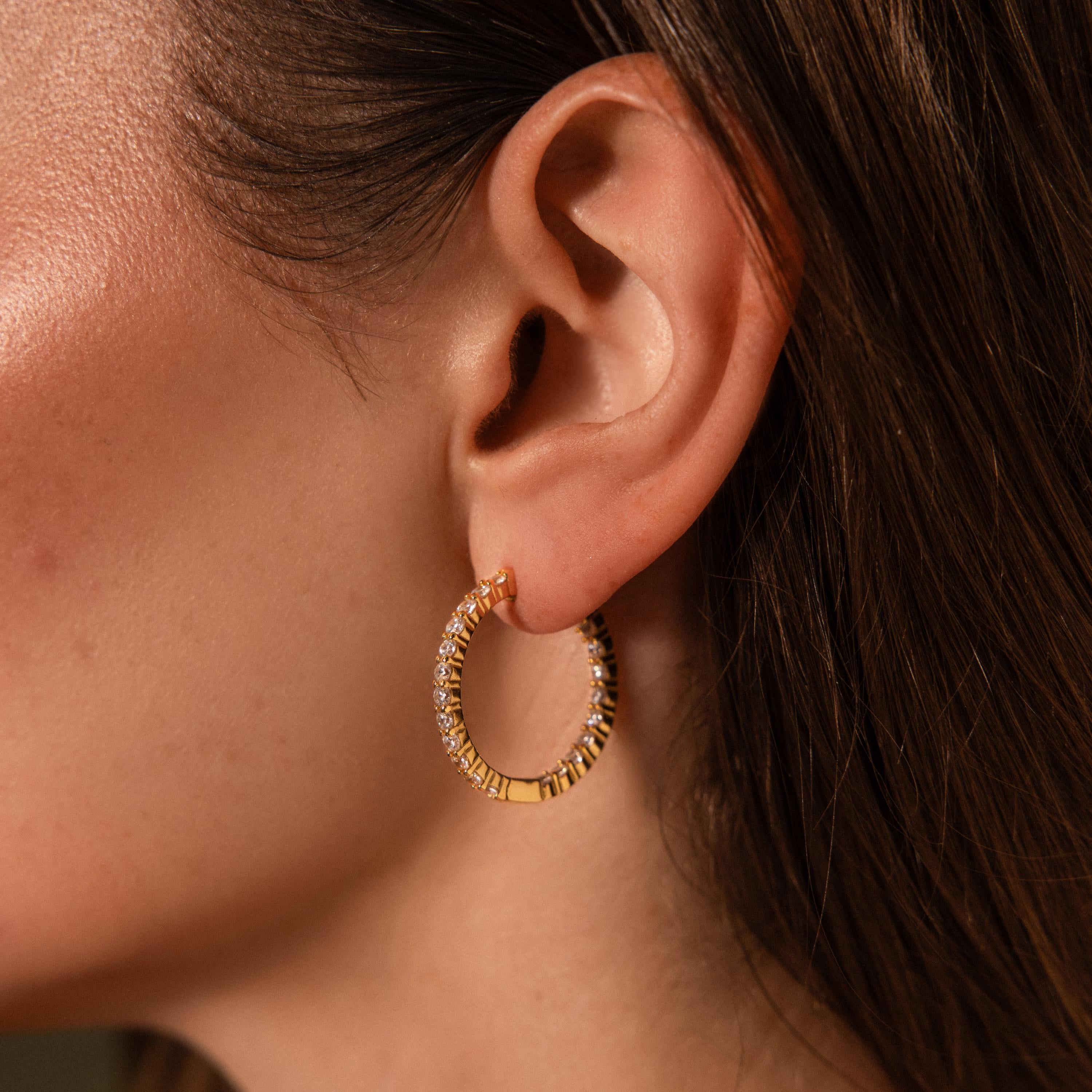 A woman’s ear is elegantly adorned with Inside Out Diamond Hoops, 18k gold hoops encrusted with sparkling gemstones.