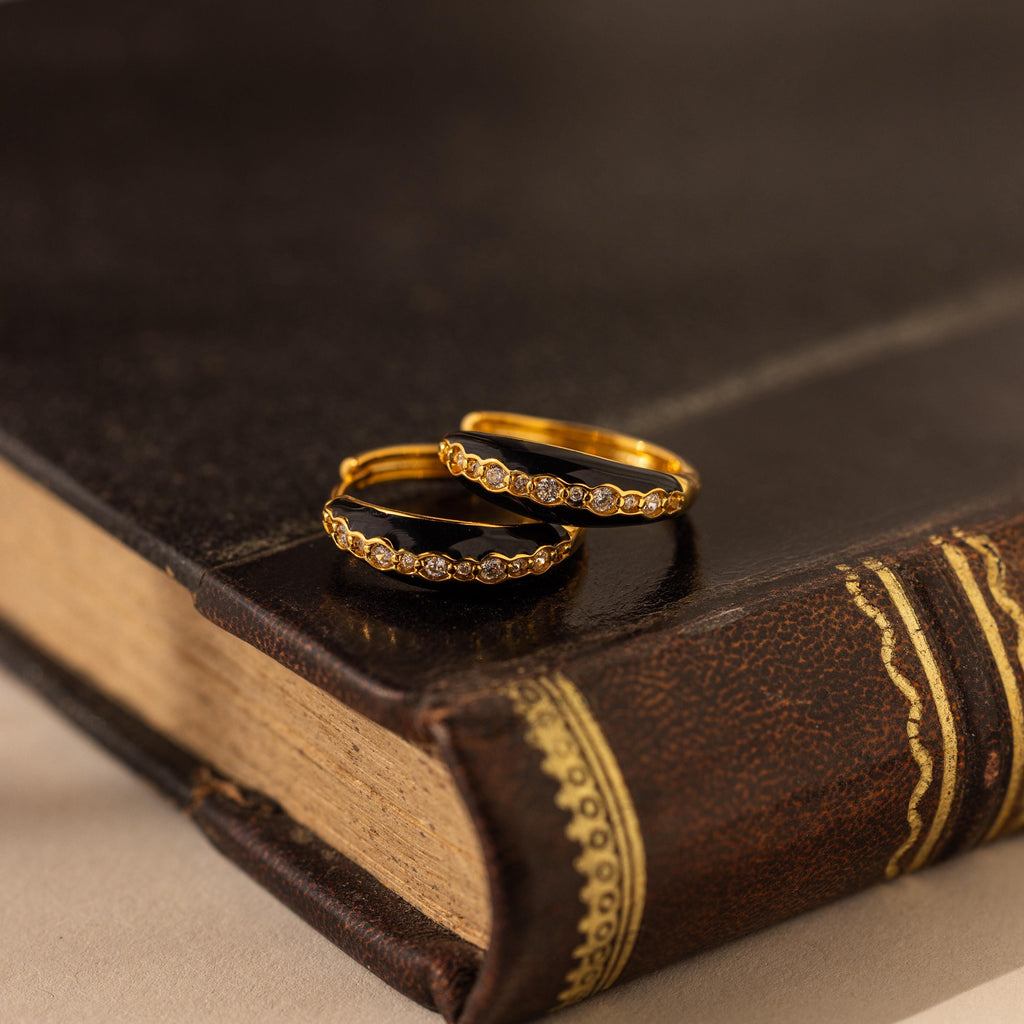 Two gold rings with small diamonds rest on a closed, vintage leather-bound book, evoking the timeless allure of Noir Pave Dome Hoops in 18K Gold.