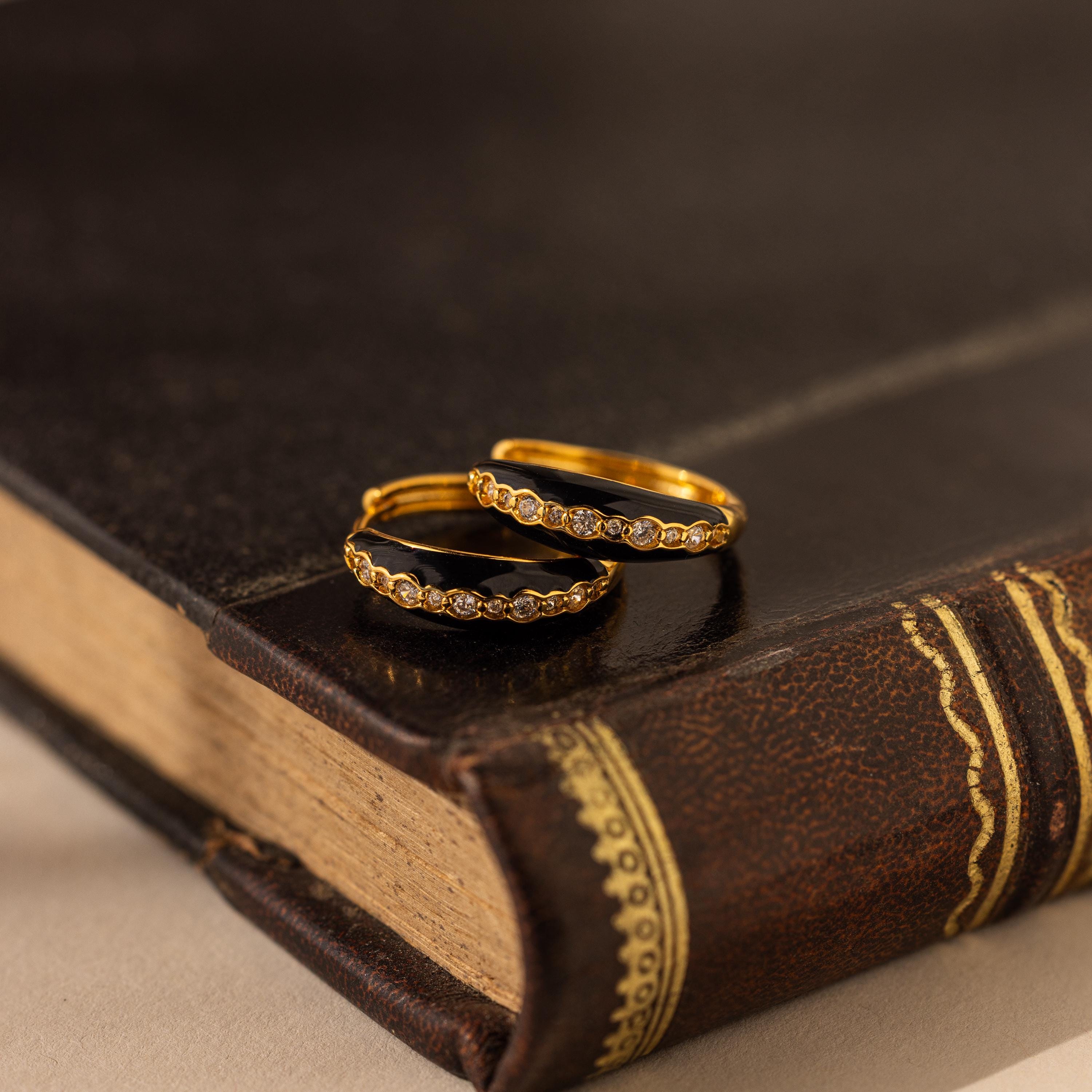 Two gold rings with small diamonds rest on a closed, vintage leather-bound book, evoking the timeless allure of Noir Pave Dome Hoops in 18K Gold.