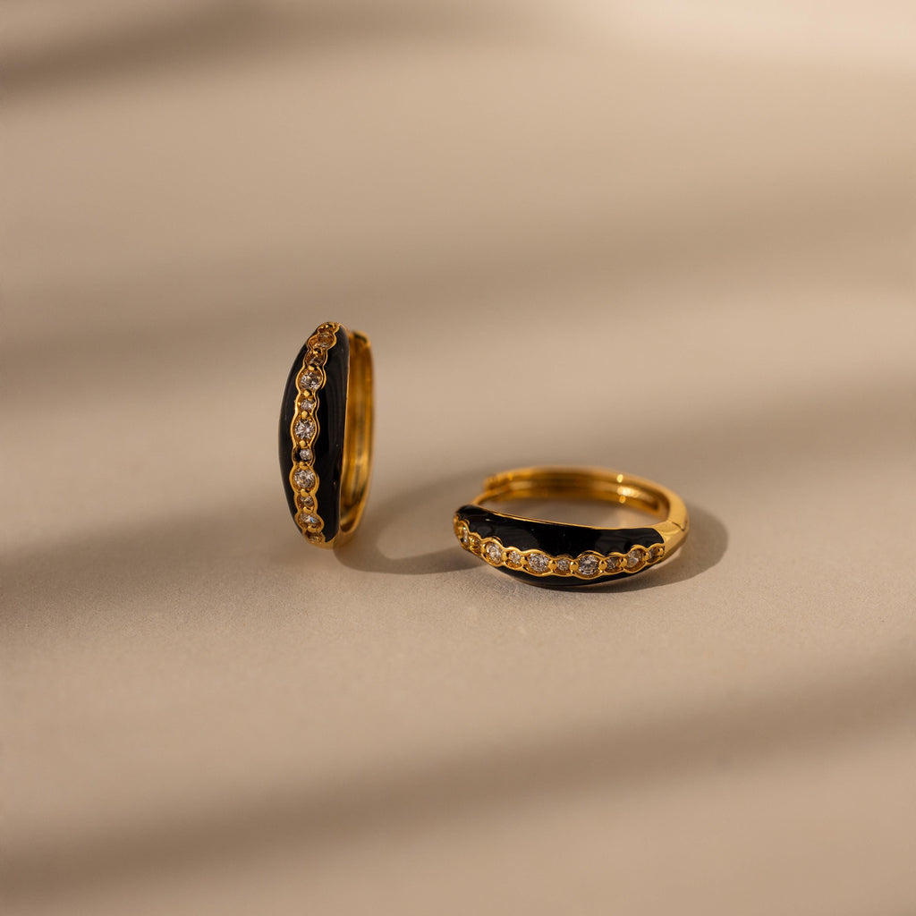 The Noir Pave Dome Hoops—gold hoop earrings featuring small diamonds and pave detailing—are displayed on a beige surface with soft shadow lines.