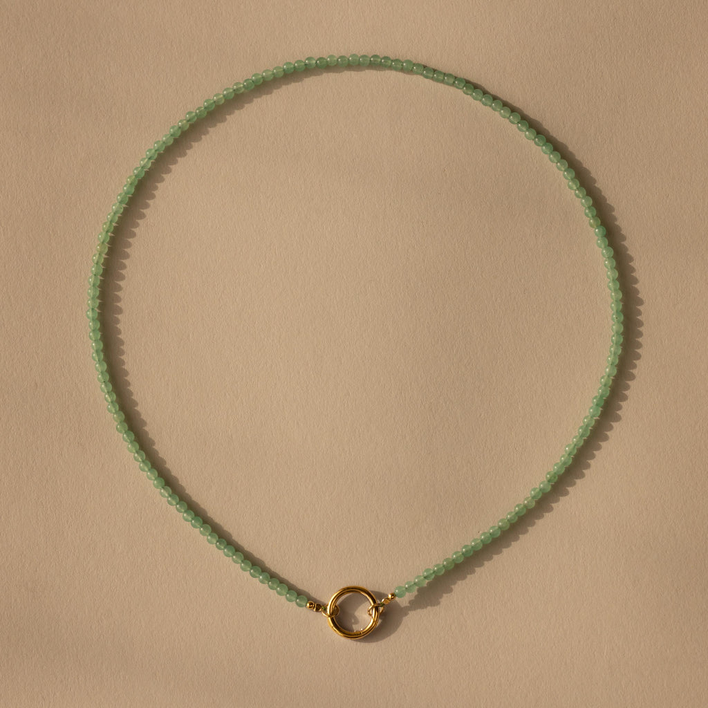 The Beaded Jade Clasp Necklace showcases smooth green jade beads and a small gold circular clasp, elegantly displayed on a beige background for a refined, sophisticated look.