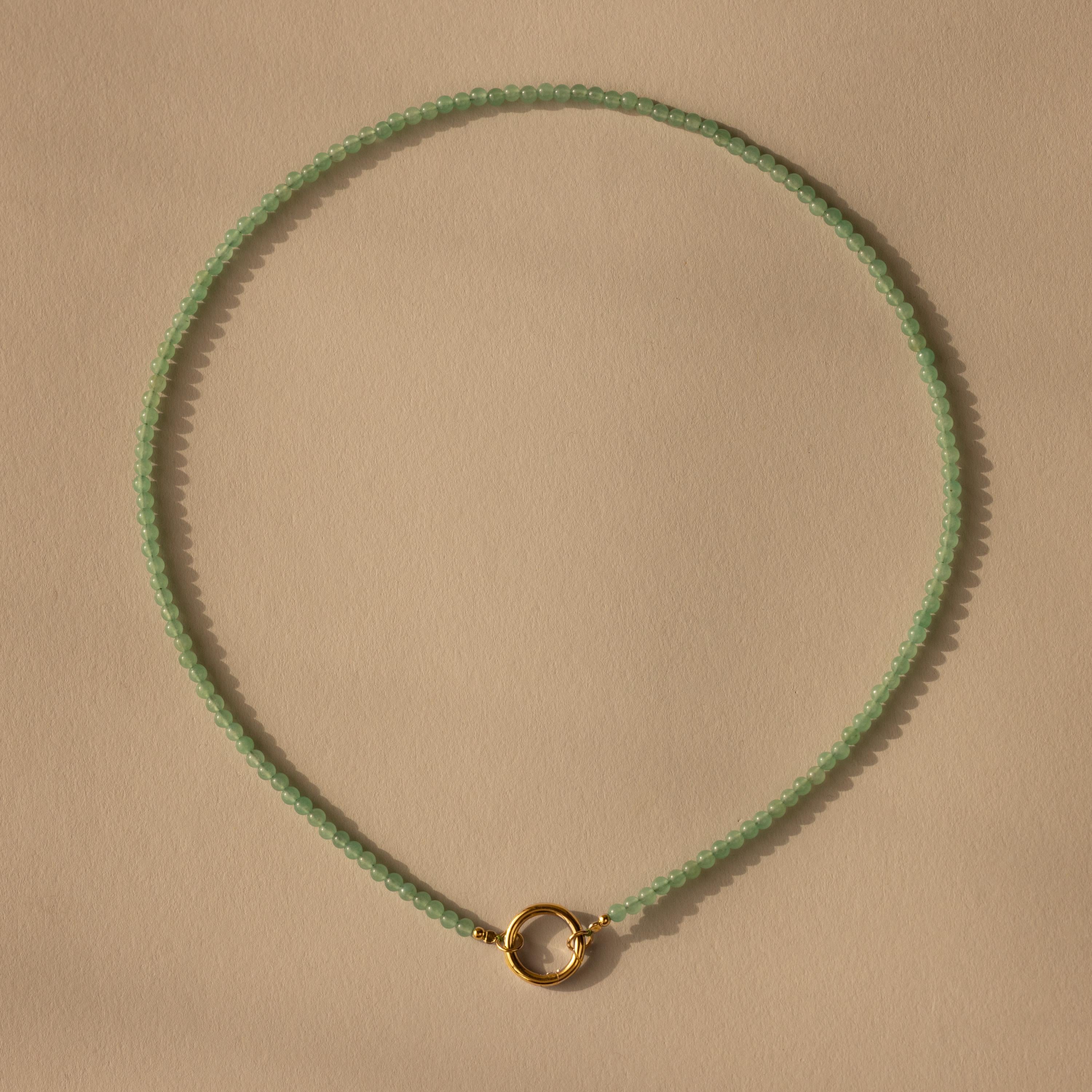 The Beaded Jade Clasp Necklace showcases smooth green jade beads and a small gold circular clasp, elegantly displayed on a beige background for a refined, sophisticated look.