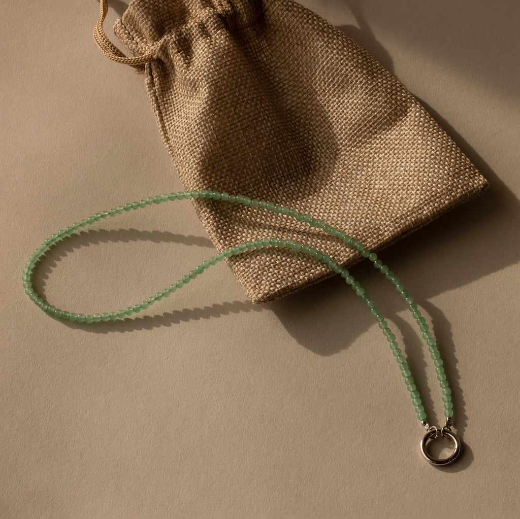 The Beaded Jade Clasp Necklace in Silver, ideal for layering, is displayed beside a brown burlap drawstring pouch on a beige surface.