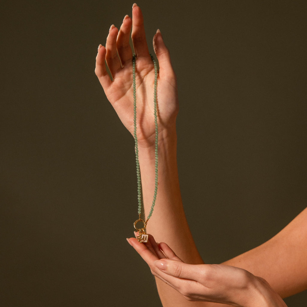 A hand displays the Beaded Jade Clasp Necklace in 18K Gold, featuring jade beads, a gold ring, and a small gold pendant—ideal for layering—set against a plain brown background.