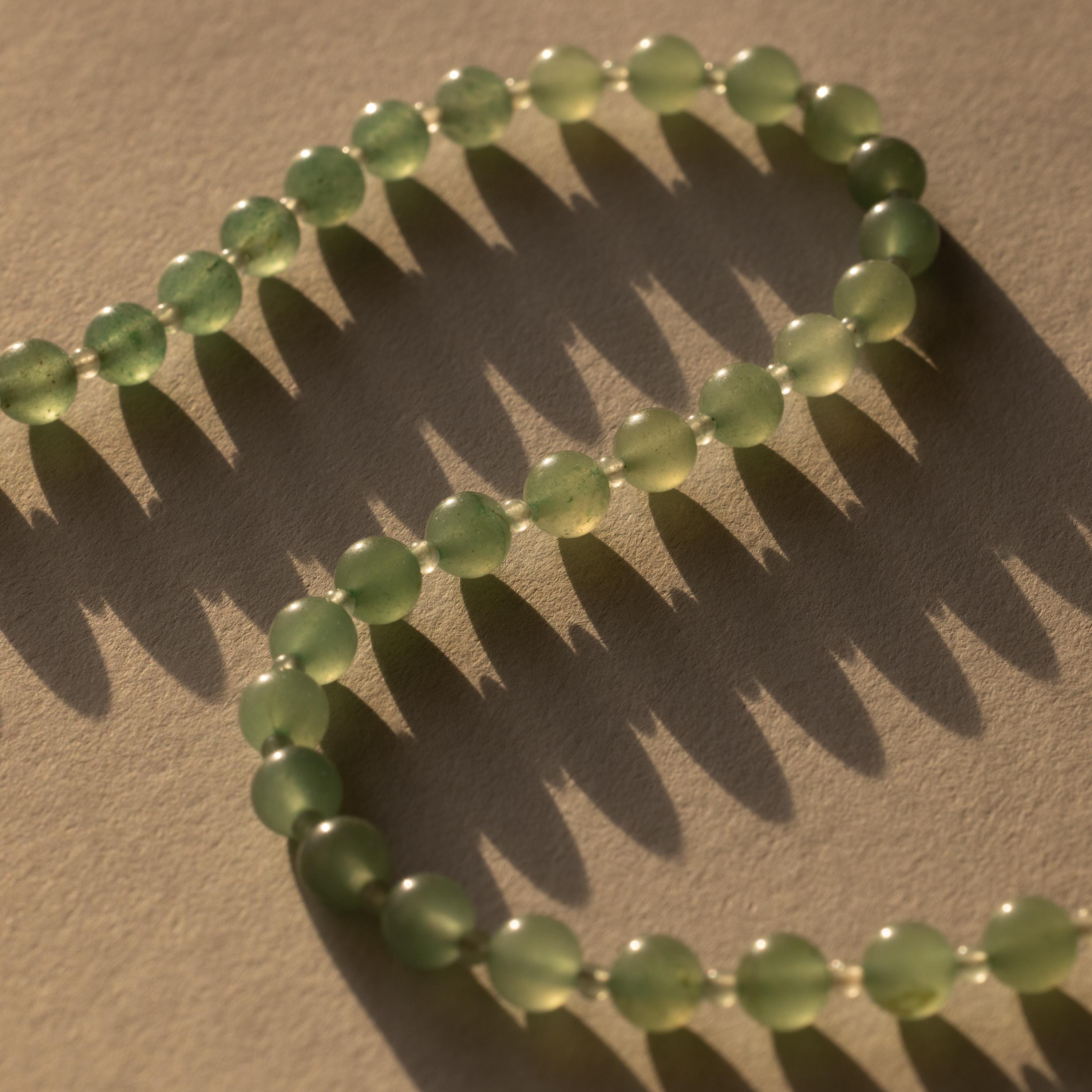 The Jade Choker Necklace in 18K Gold features green jade beads that cast soft, patterned shadows on a beige textured surface under warm sunlight.
