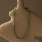The Jade Choker Necklace with green beads is shown on a textured, abstract beige bust set against a neutral background.