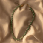 The Jade Choker Necklace, featuring green beads and a gold clasp, is displayed on soft beige fabric with gentle shadows.