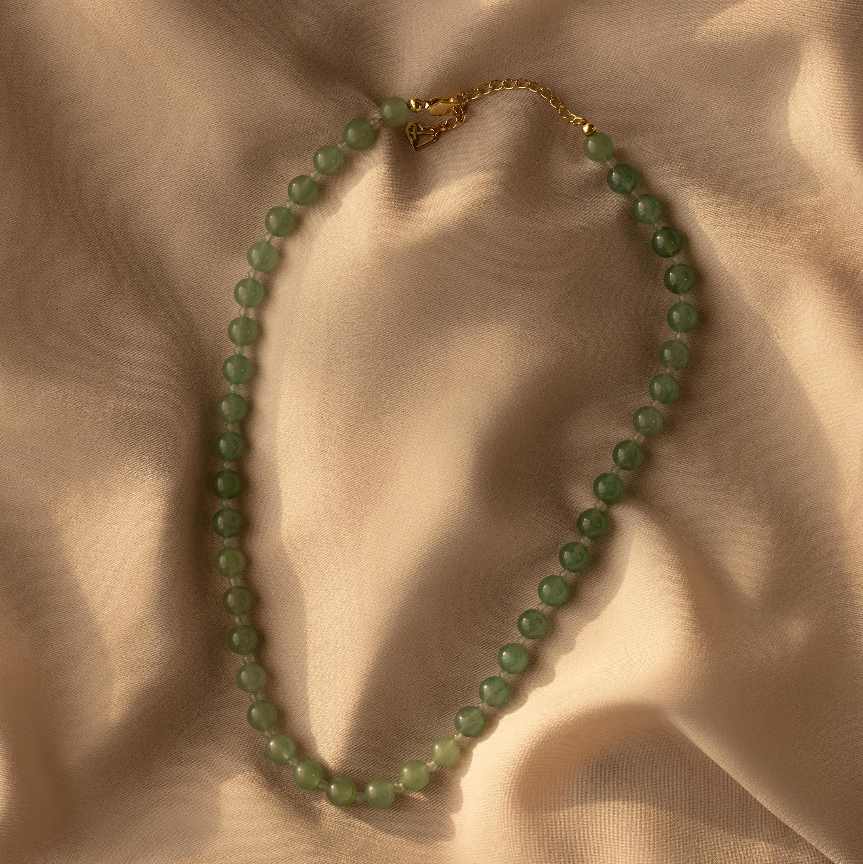 The Jade Choker Necklace, featuring green beads and a gold clasp, is displayed on soft beige fabric with gentle shadows.