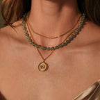 A woman wears layered necklaces: a Jade Choker Necklace in 18K Gold, a gold chain, and a gold pendant necklace.