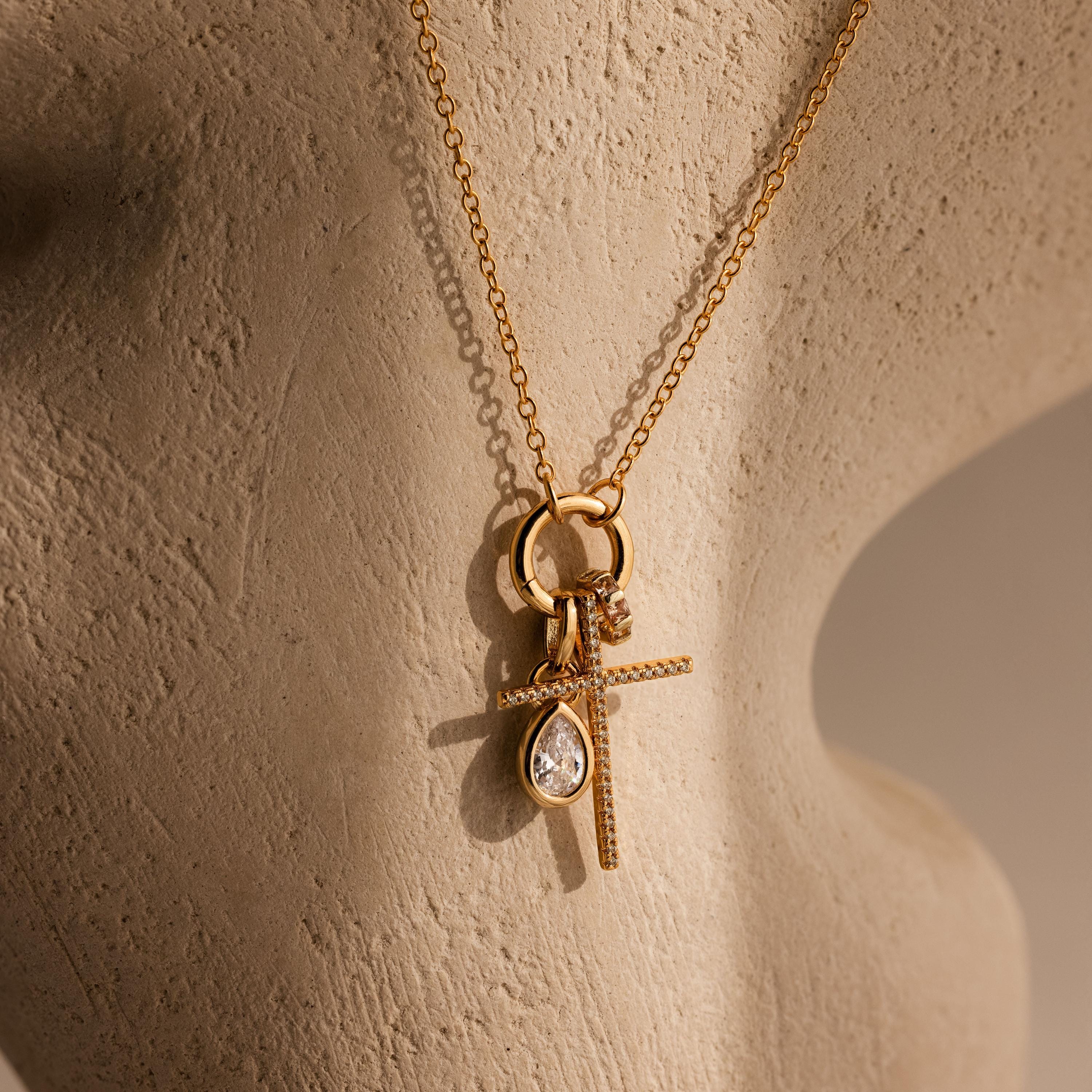 The Skinny Pave Cross Charm in 18K Gold, paired with a teardrop gemstone pendant and displayed on a beige sculpted mannequin, is perfect for those seeking elegant faith jewelry.