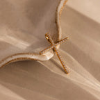 The Skinny Pave Cross Charm, featuring petite diamonds, rests on a textured beige ceramic surface and casts soft shadows—a timeless symbol of faith.