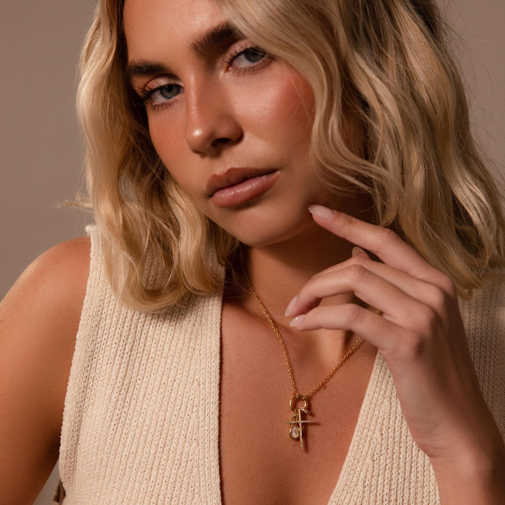A woman with wavy blonde hair wears a cream knit top and the Skinny Pave Cross Charm necklace, posing thoughtfully.