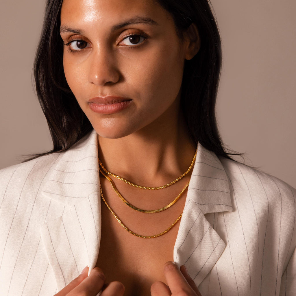 A woman in a white pinstripe blazer layers the Twisted Snake Chain Necklace with two gold chains, styled against a neutral background.