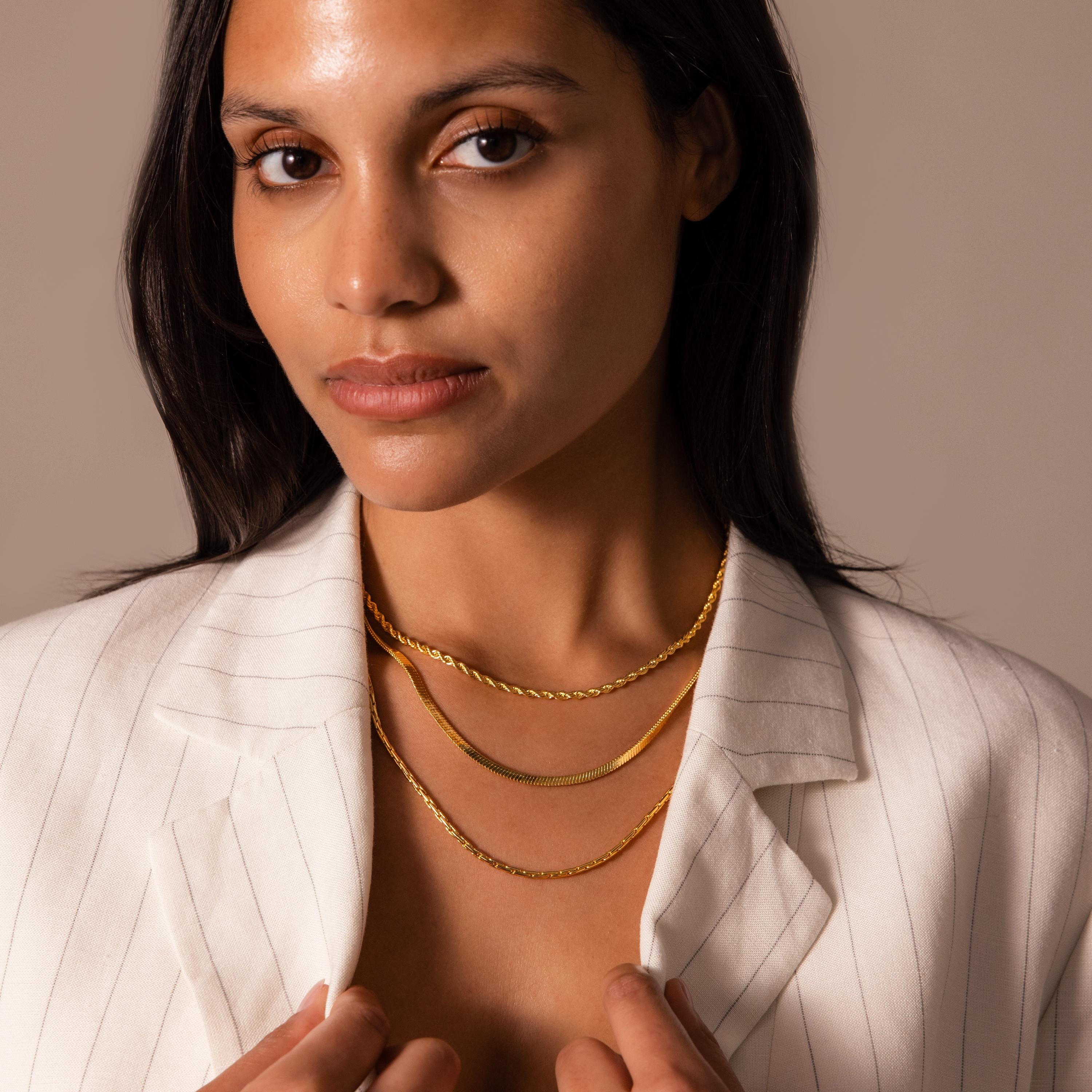 A woman in a white pinstripe blazer layers the Twisted Snake Chain Necklace with two gold chains, styled against a neutral background.