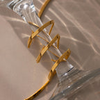 The Twisted Snake Chain Necklace in 18K Gold is wrapped around the stem of a clear glass, casting soft shadows on a beige surface.