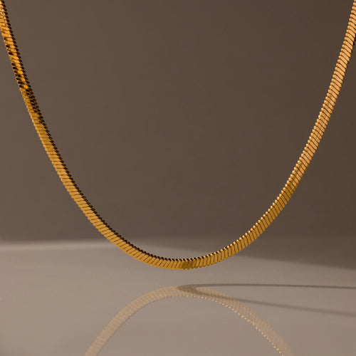 The Bold Snake Chain Necklace in 18K Gold is showcased on a neutral brown background with a soft shadow beneath, highlighting its modern design.