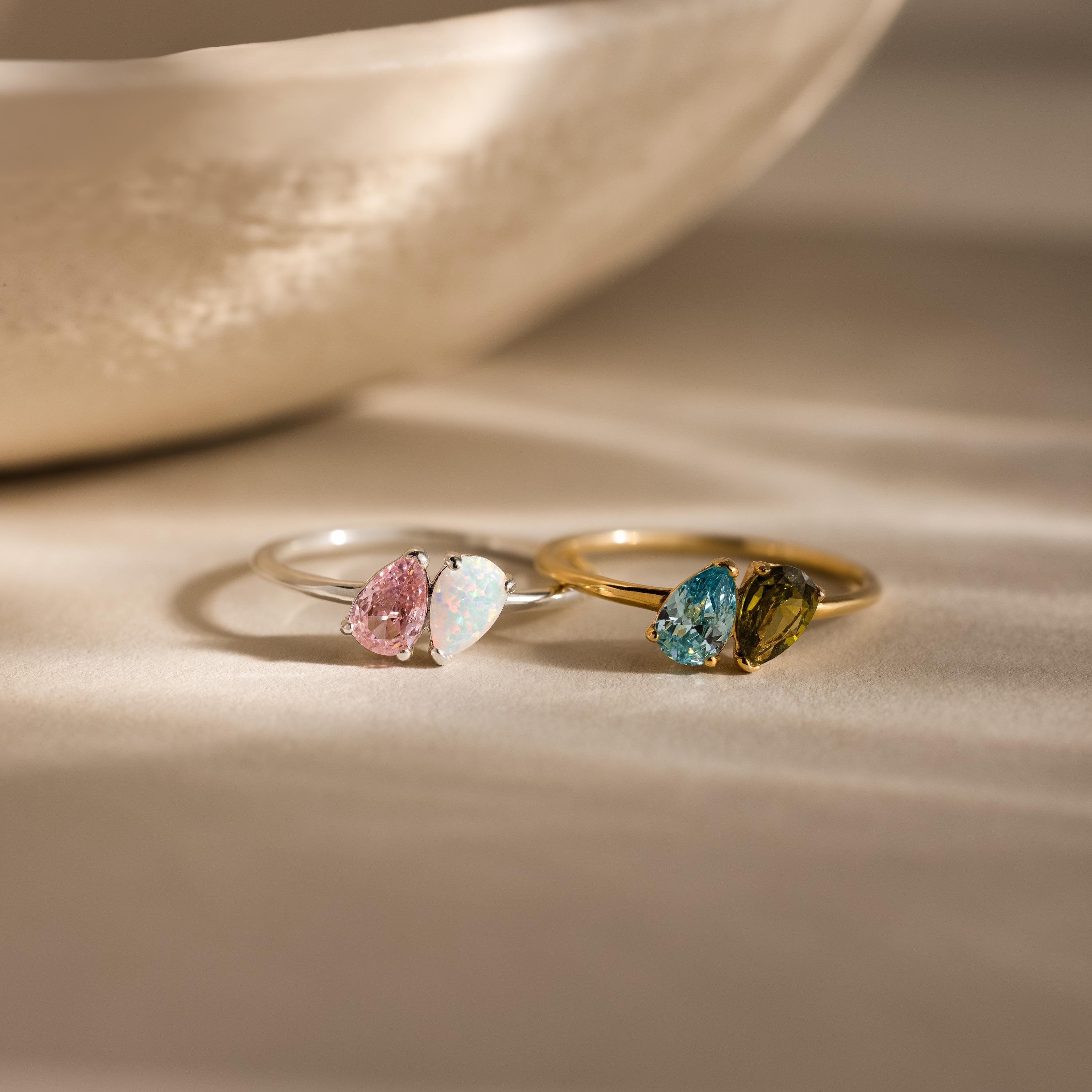 The Duo Pear Toi Et Moi Ring features two elegant pear-shaped stones—one silver with pink and clear gems, the other gold with blue and green—adding romantic charm to any look.