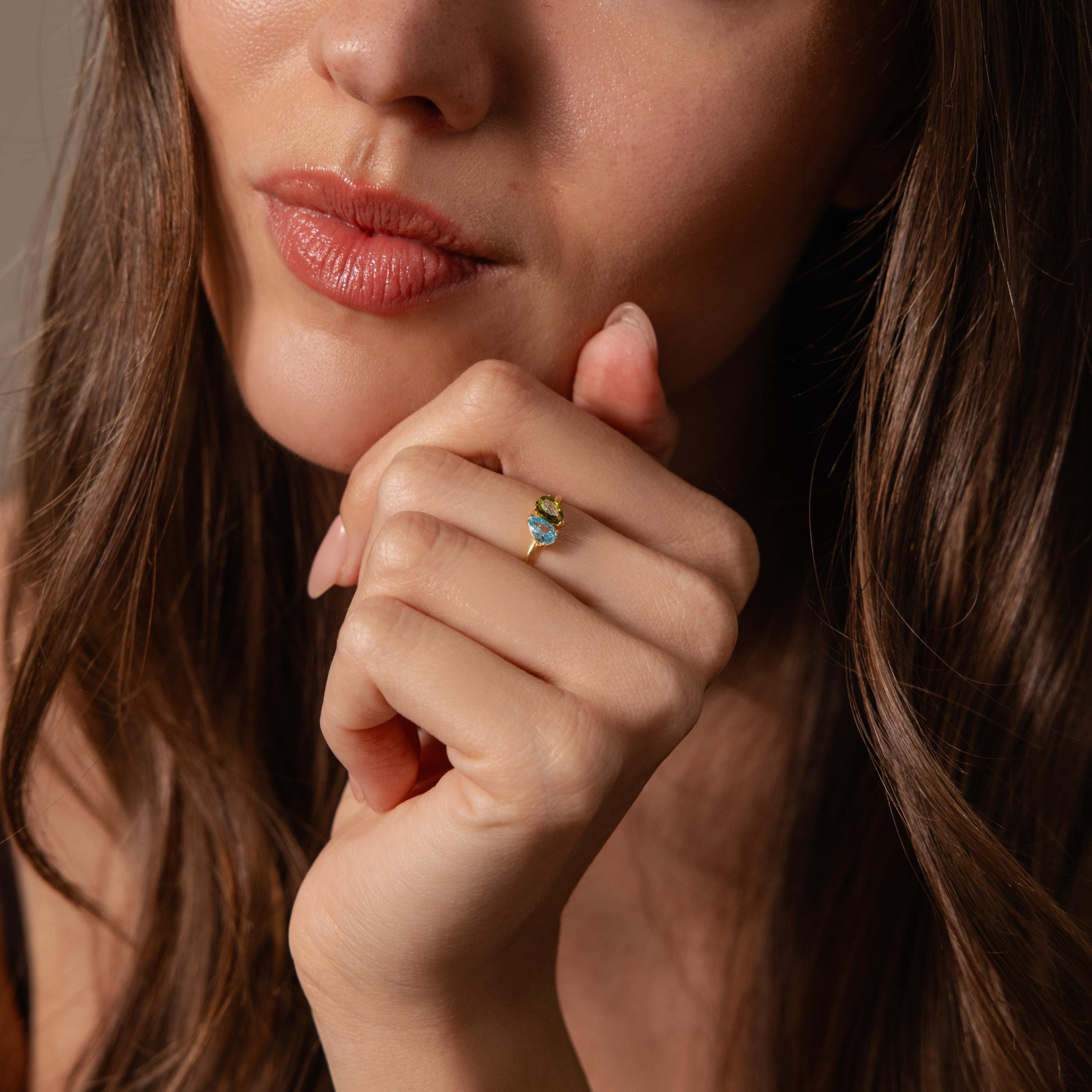 A woman with long brown hair rests her chin on her hand, wearing the Duo Pear Toi Et Moi Ring, a romantic piece featuring elegant green pear-shaped stones.