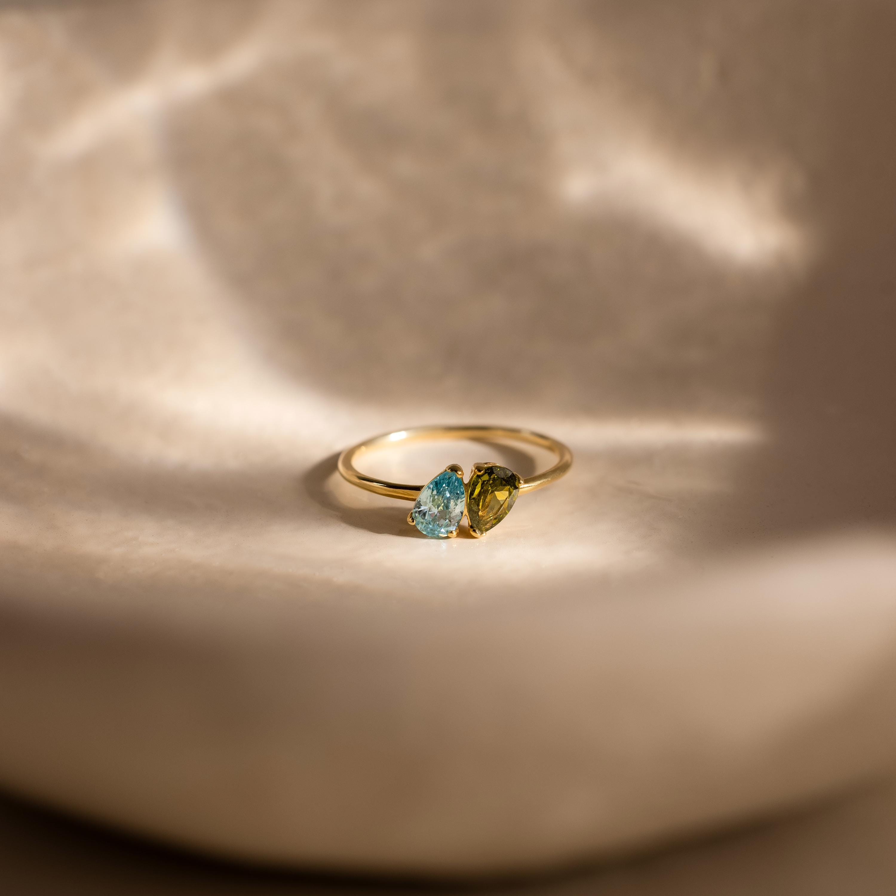 The Duo Pear Toi Et Moi Ring, featuring blue and green pear-shaped gemstones, shines on a softly lit beige surface—a romantic piece.