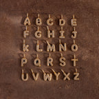 Pave Bubble Letter Charms featuring diamond-studded alphabet designs are displayed in four rows on a brown textured surface.
