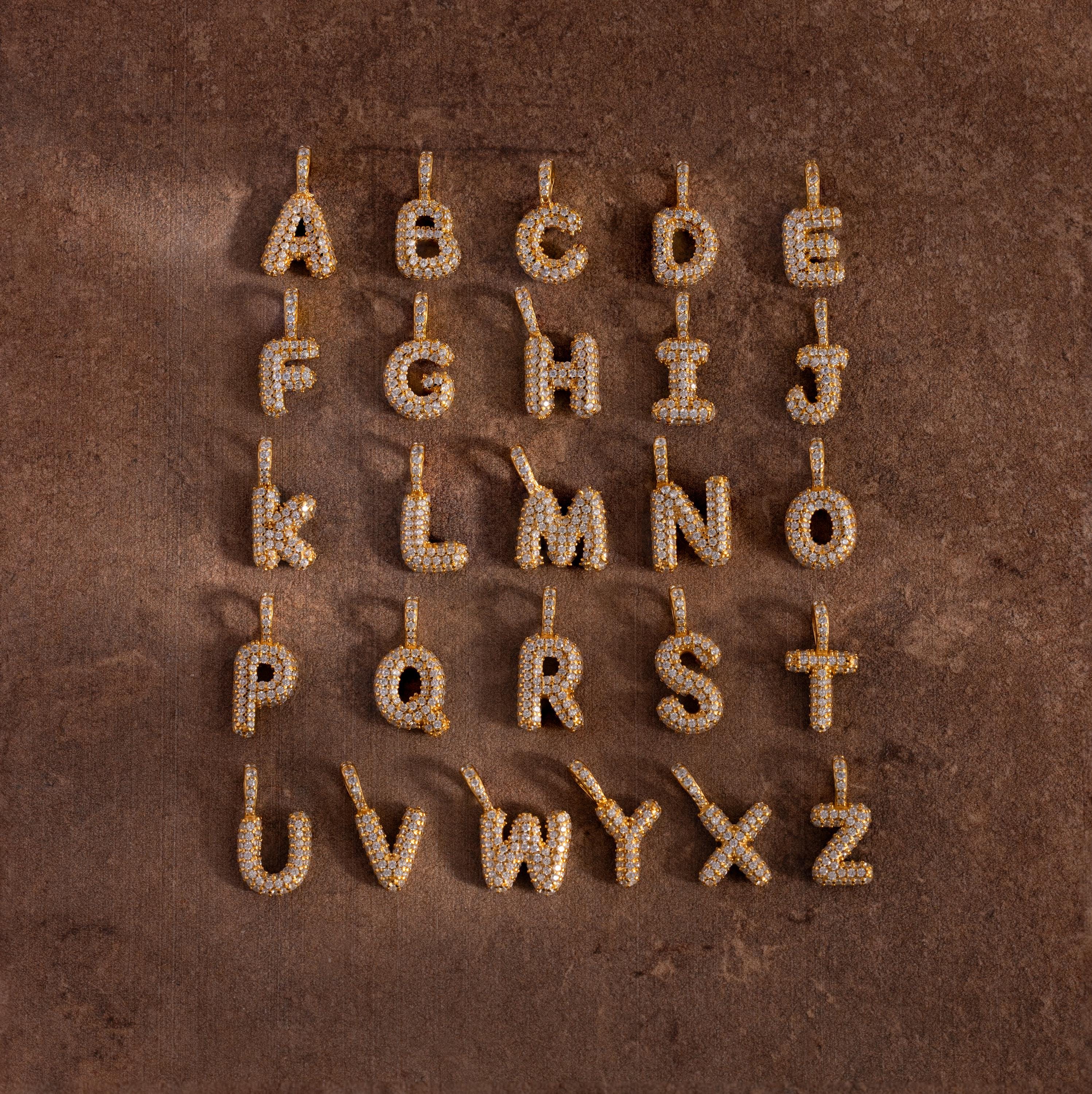 Pave Bubble Letter Charms featuring diamond-studded alphabet designs are displayed in four rows on a brown textured surface.