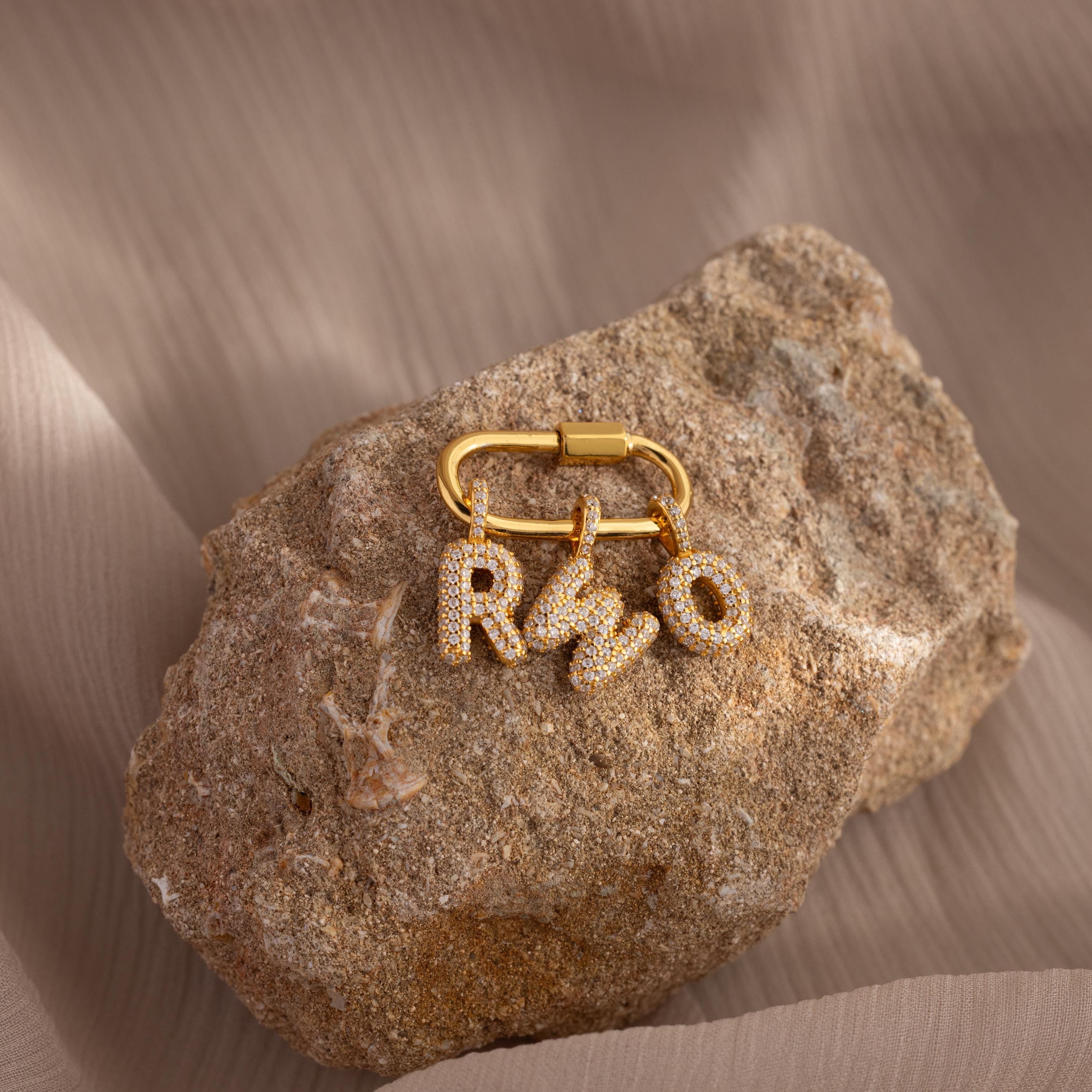 A gold keyring with Pave Bubble Letter Charms, each charm elegantly studded with diamonds in the letters R, S, and O, rests on a textured rock.