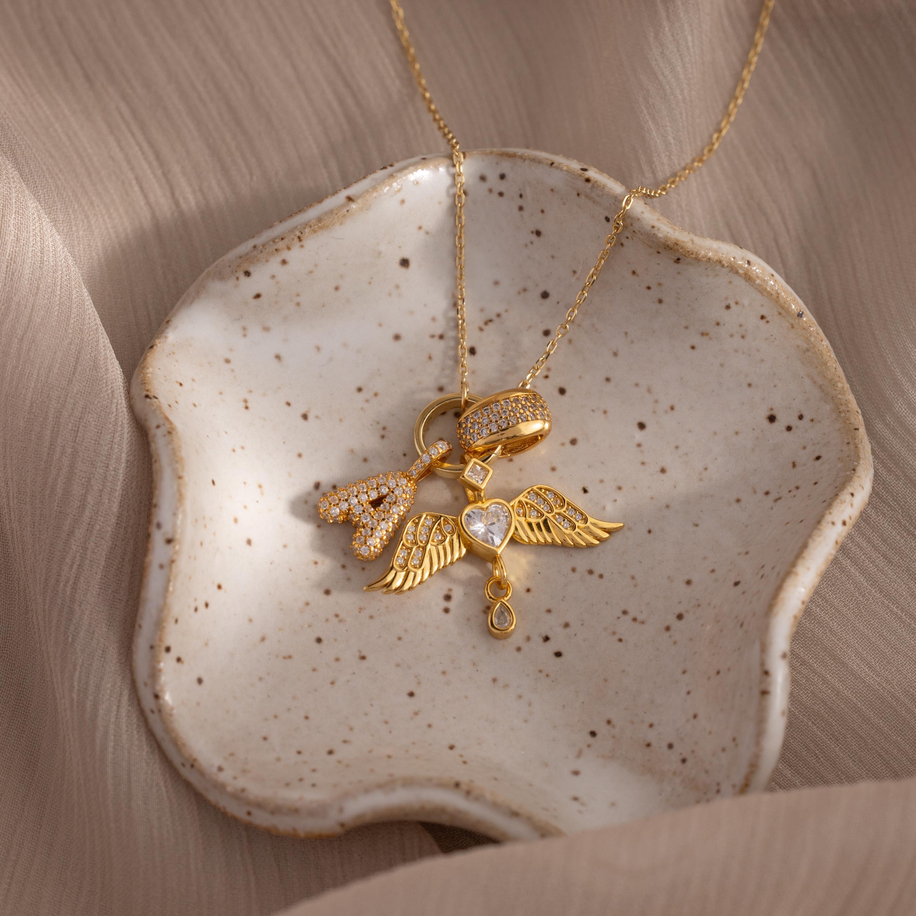 The Pave Bubble Letter Charm gold necklace with heart and wing rests on a speckled ceramic dish, set against a beige fabric background.
