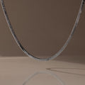 The Twisted Snake Chain Necklace in Silver hangs gracefully, its reflection shimmering on a smooth surface—an ideal choice for fans of modern jewelry.