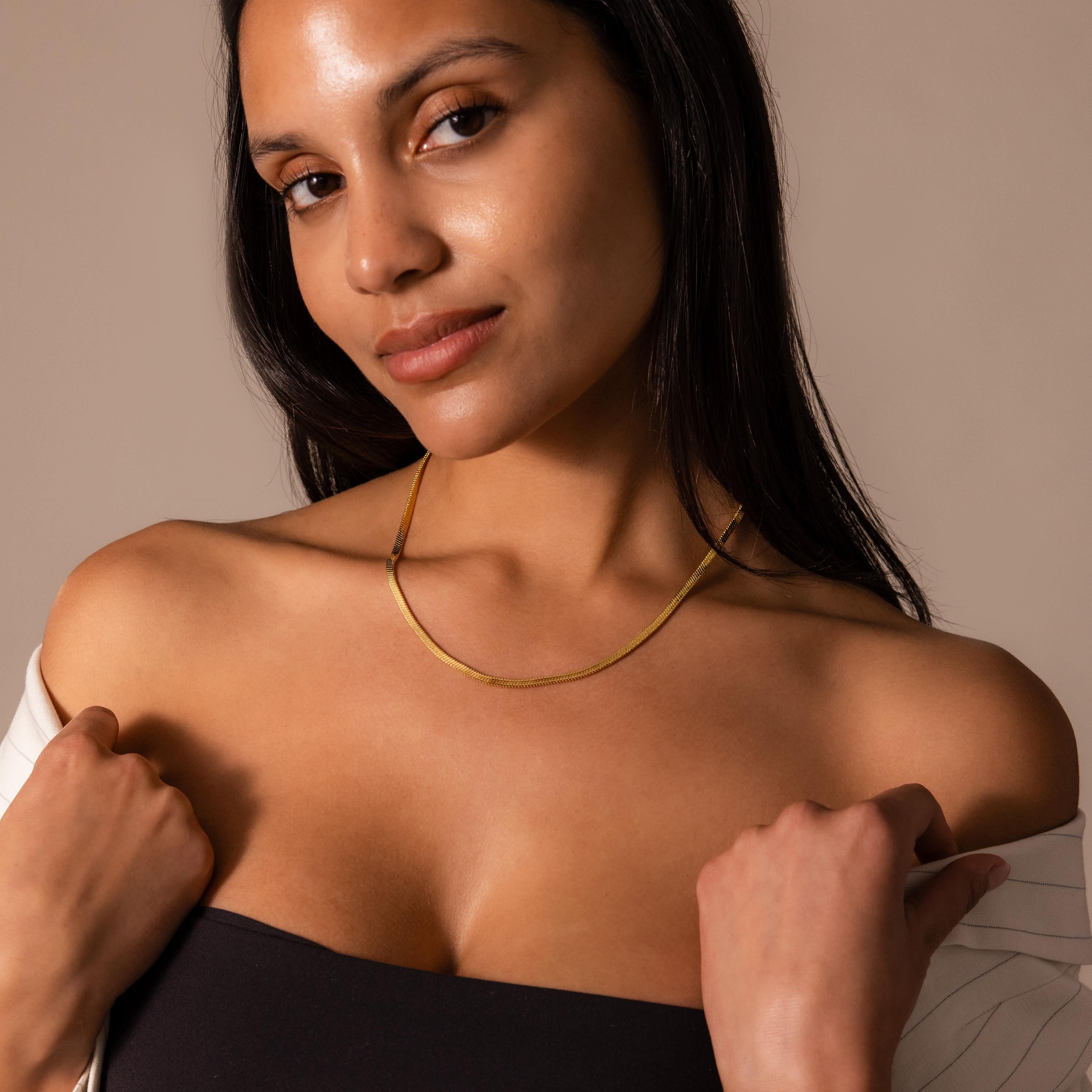 Woman with long dark hair wears a Twisted Snake Chain Necklace in 18K Gold and an off-the-shoulder top, posing confidently to showcase her modern jewelry style.
