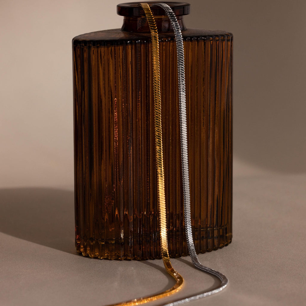 The Bold Snake Chain Necklace, accented with gold and silver chains, is elegantly displayed on an amber glass bottle against a neutral background.