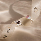 Two Duo Pear Toi Et Moi Rings in Silver, featuring colorful pear-shaped stones, are placed on a cream ribbon with gold lettering and a heart logo, creating a romantic display.