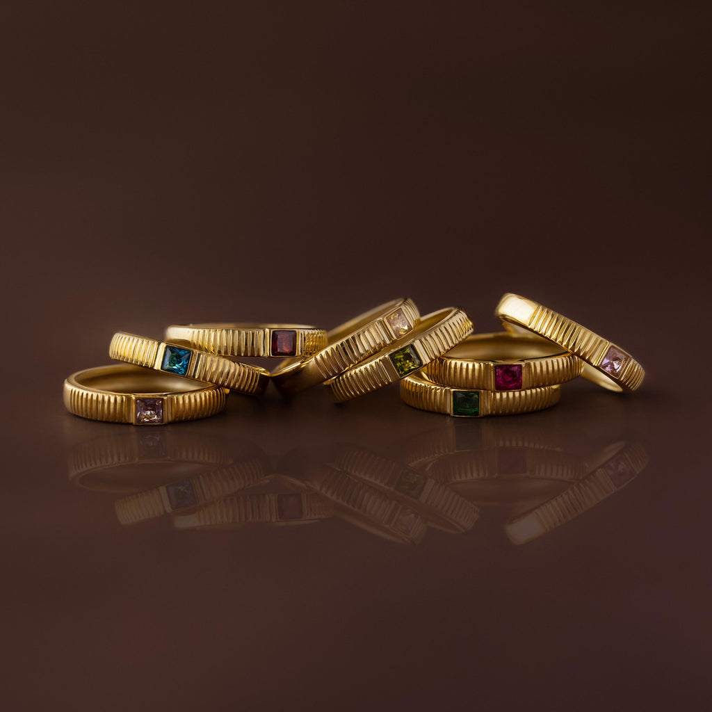 Seven gold rings, including the Ribbed Pink Tourmaline Band, showcase vibrant gemstones and are artfully grouped on a glossy brown surface.