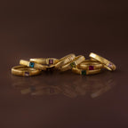 Seven gold rings, including the Ribbed Pink Tourmaline Band, showcase vibrant gemstones and are artfully grouped on a glossy brown surface.