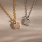 The Square Mother of Pearl Necklace, featuring an iridescent square pendant, rests on a beige textured surface—ideal for those who love delicate jewelry.