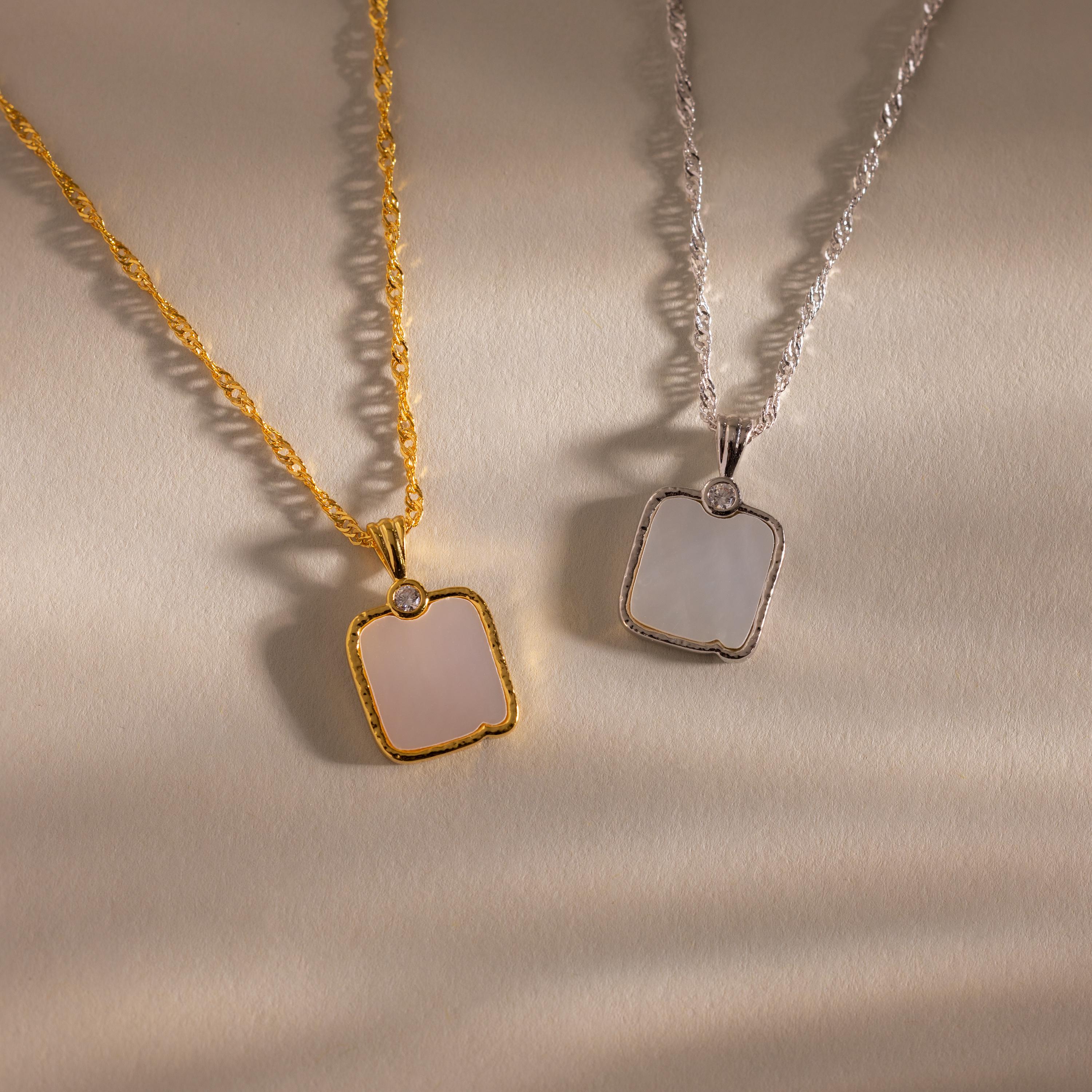 The Square Mother of Pearl Necklace, featuring an iridescent square pendant, rests on a beige textured surface—ideal for those who love delicate jewelry.