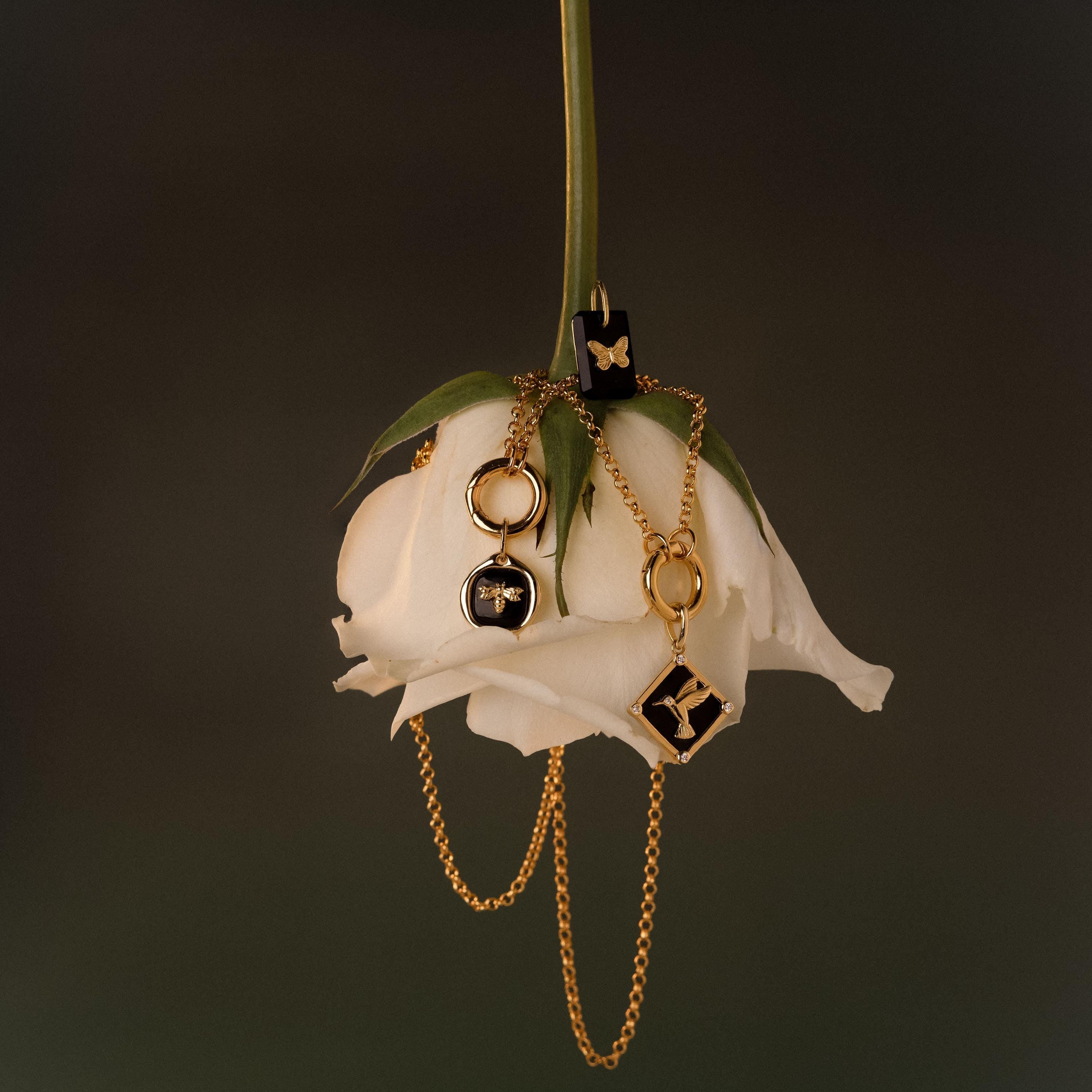 The Cable Circle Charm Necklace in 18K Gold, featuring striking black accents and a statement charm, drapes elegantly over an upside-down white rose against a dark backdrop.