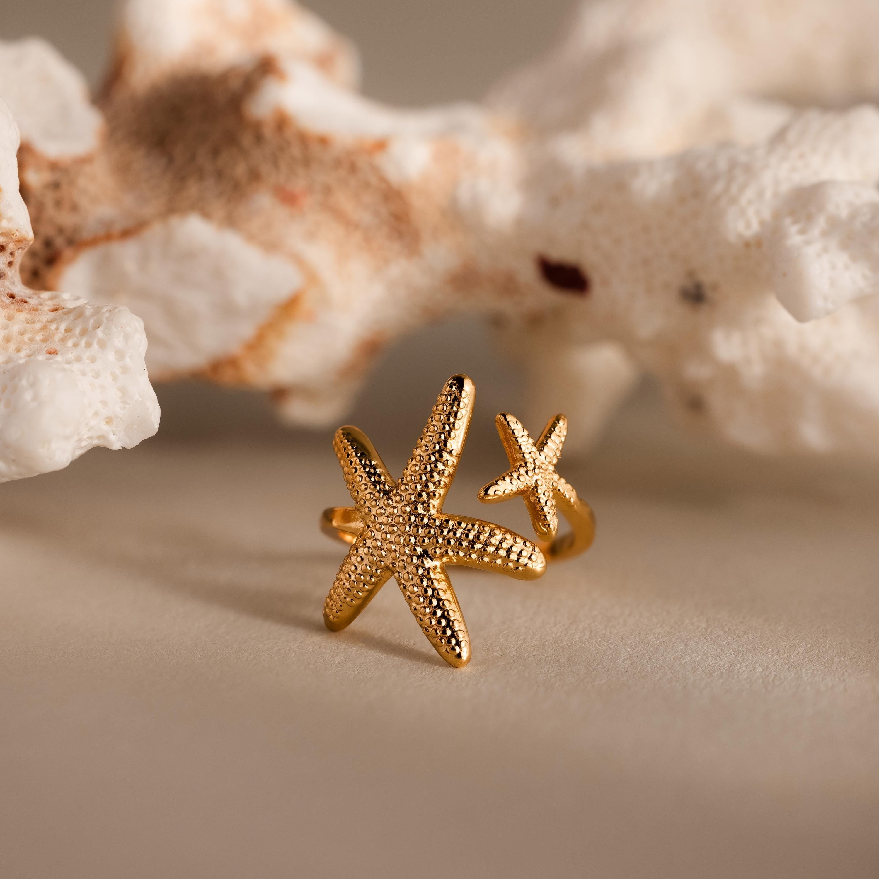 The Open Duo Starfish Ring sits on a beige surface with white coral behind it, capturing coastal charm and making a perfect statement piece.
