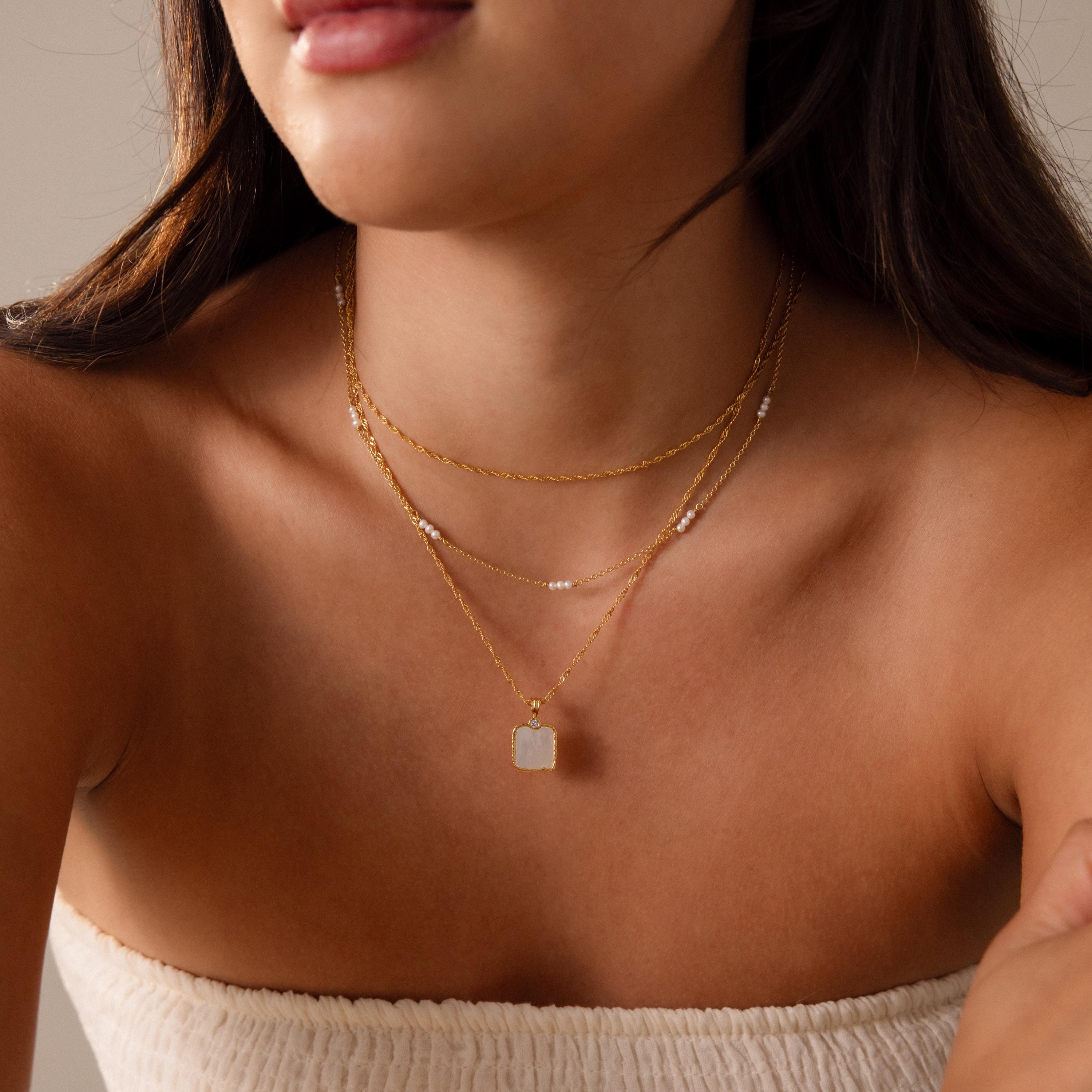 A woman wears a Square Mother of Pearl Necklace layered with gold necklaces and a white textured strapless top, soft lighting highlighting the delicate jewelry.