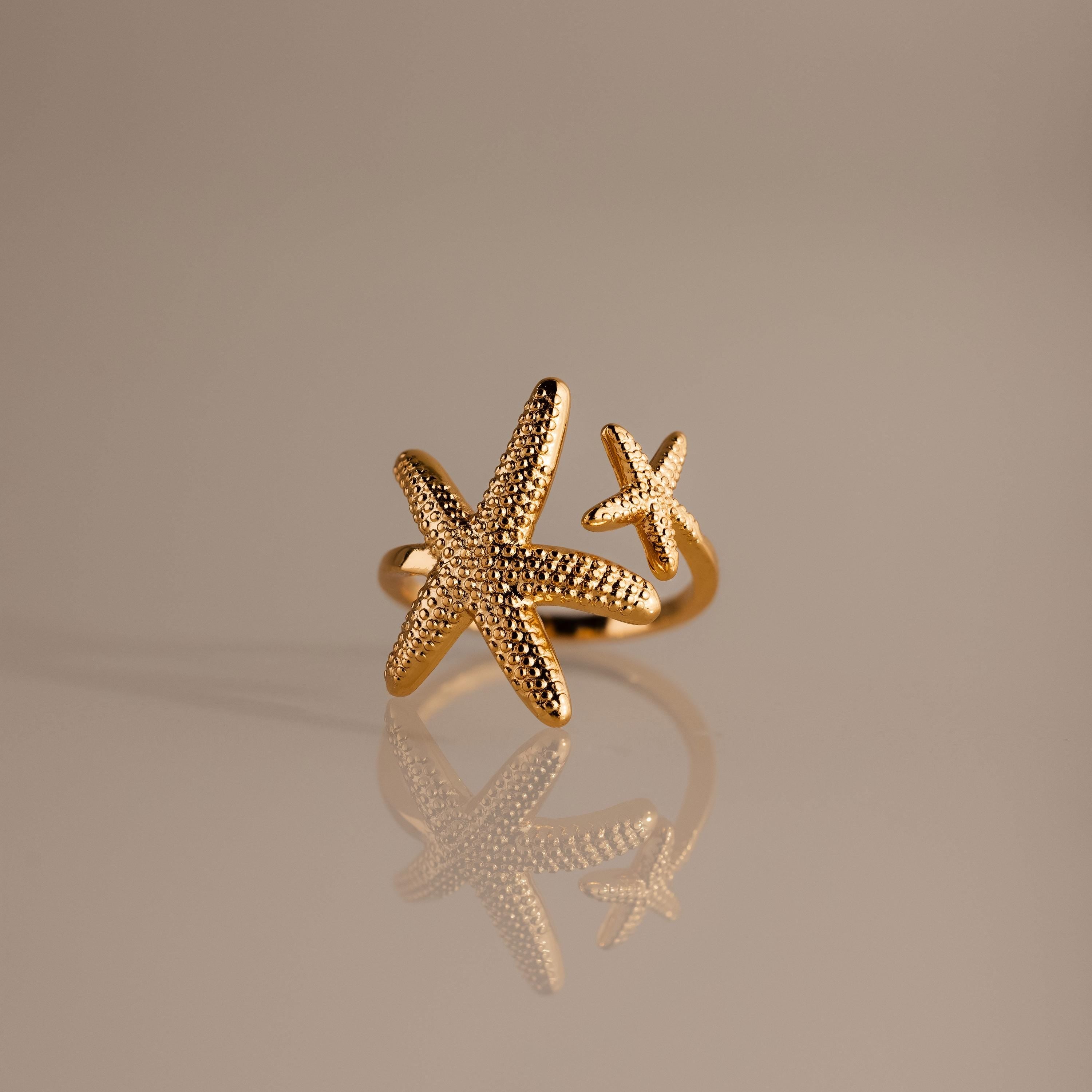 The Open Duo Starfish Ring showcases two textured starfish, one large and one small, on a reflective beige surface—a stunning gold coastal-inspired statement jewelry piece.